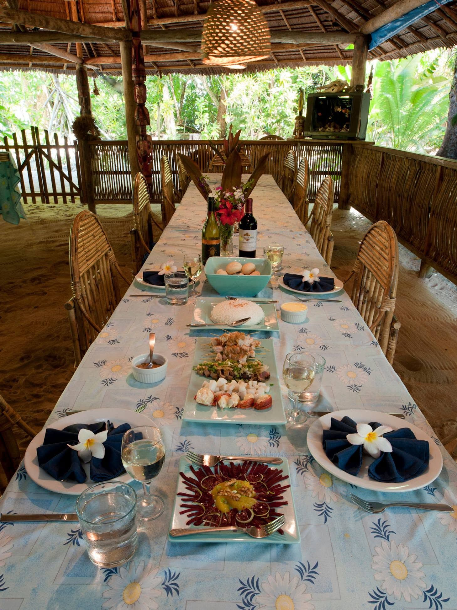 A long dining table set for a meal under a thatched roof with views of greenery.