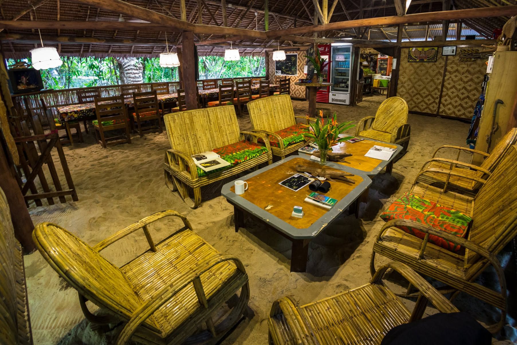 Open-air lounge area with bamboo furniture and a sandy floor under a high roof.