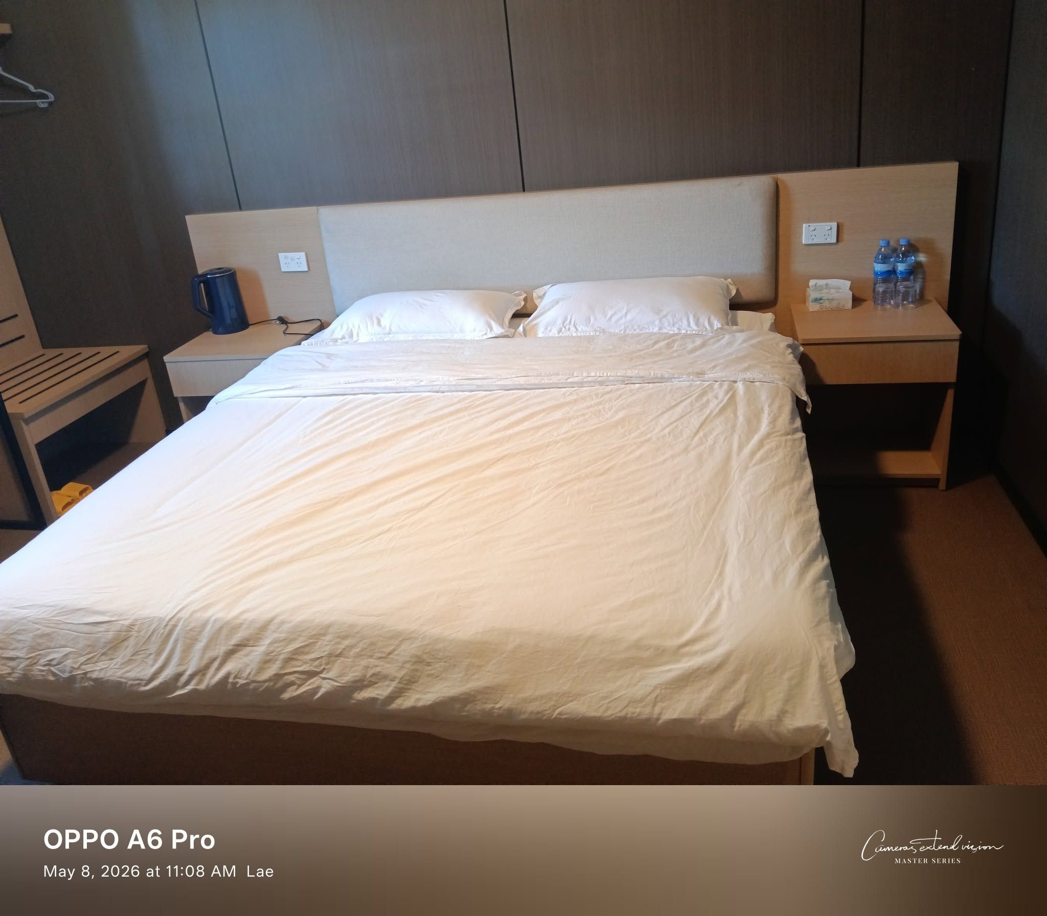Neatly made king bed flanked by wooden nightstands with bottled water and kettle.