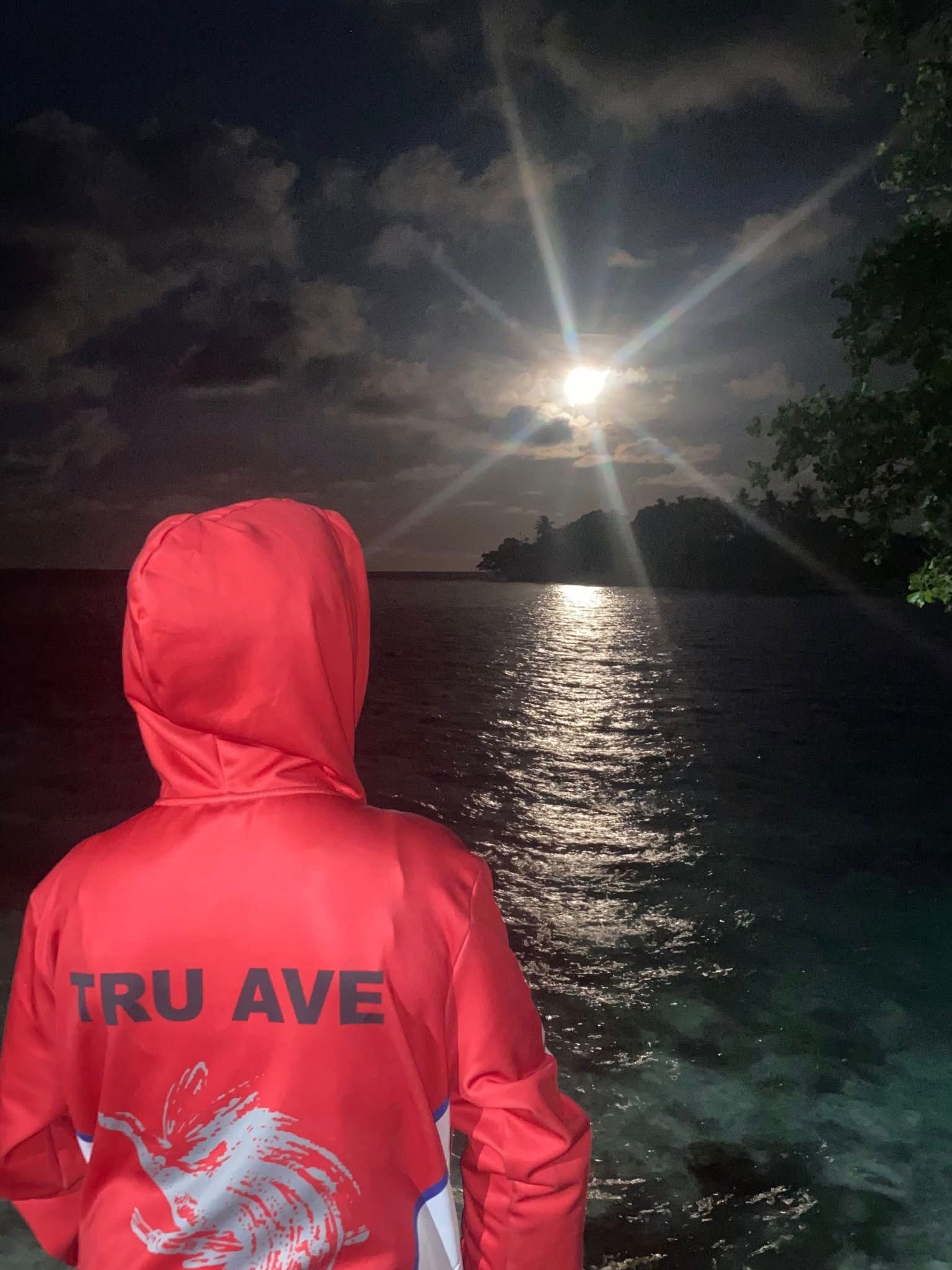 A person in a red hoodie watching the moon reflect on the water.