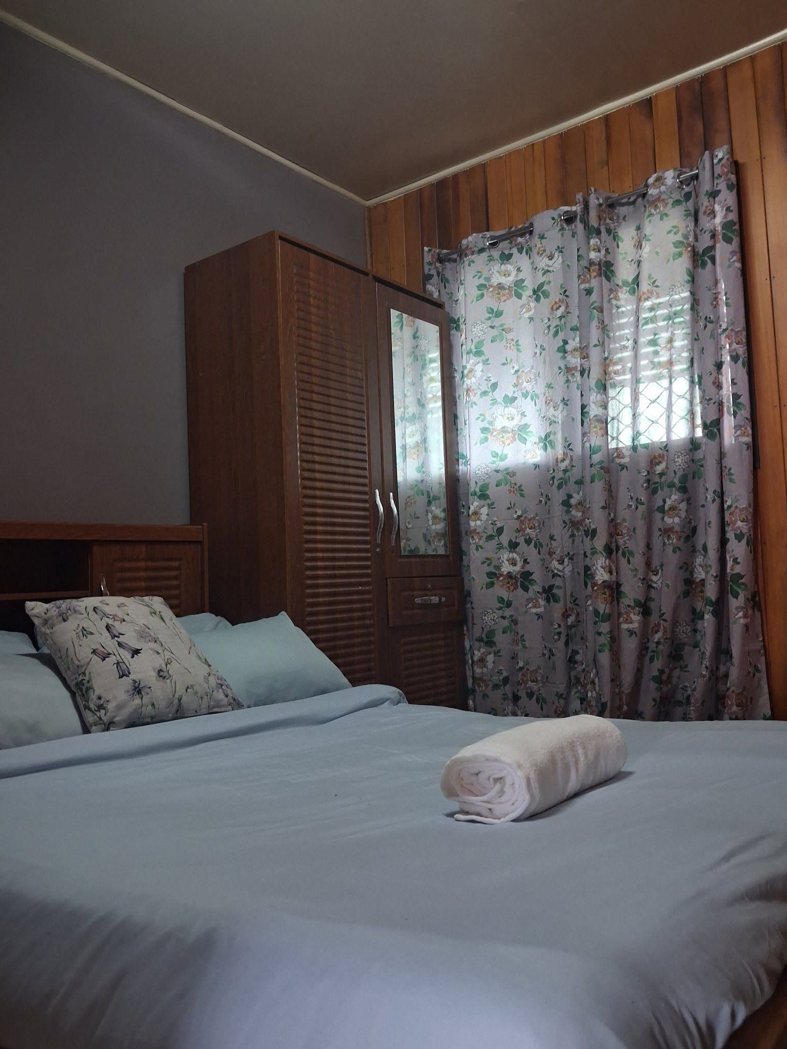 A tidy double bed with floral curtains and a wooden wardrobe.