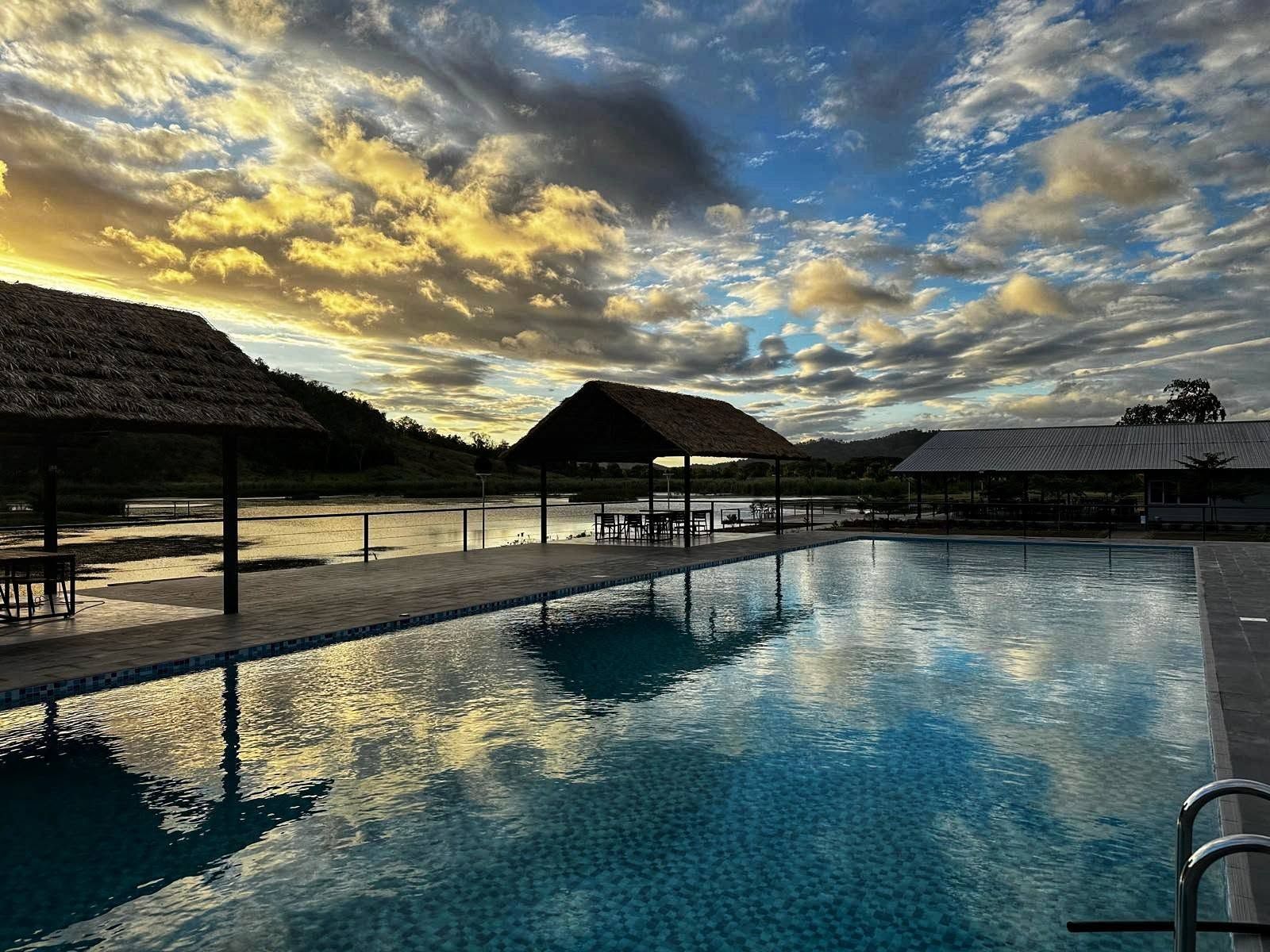 Outdoor swimming pool with thatched-roof gazebos and sunset sky reflections.