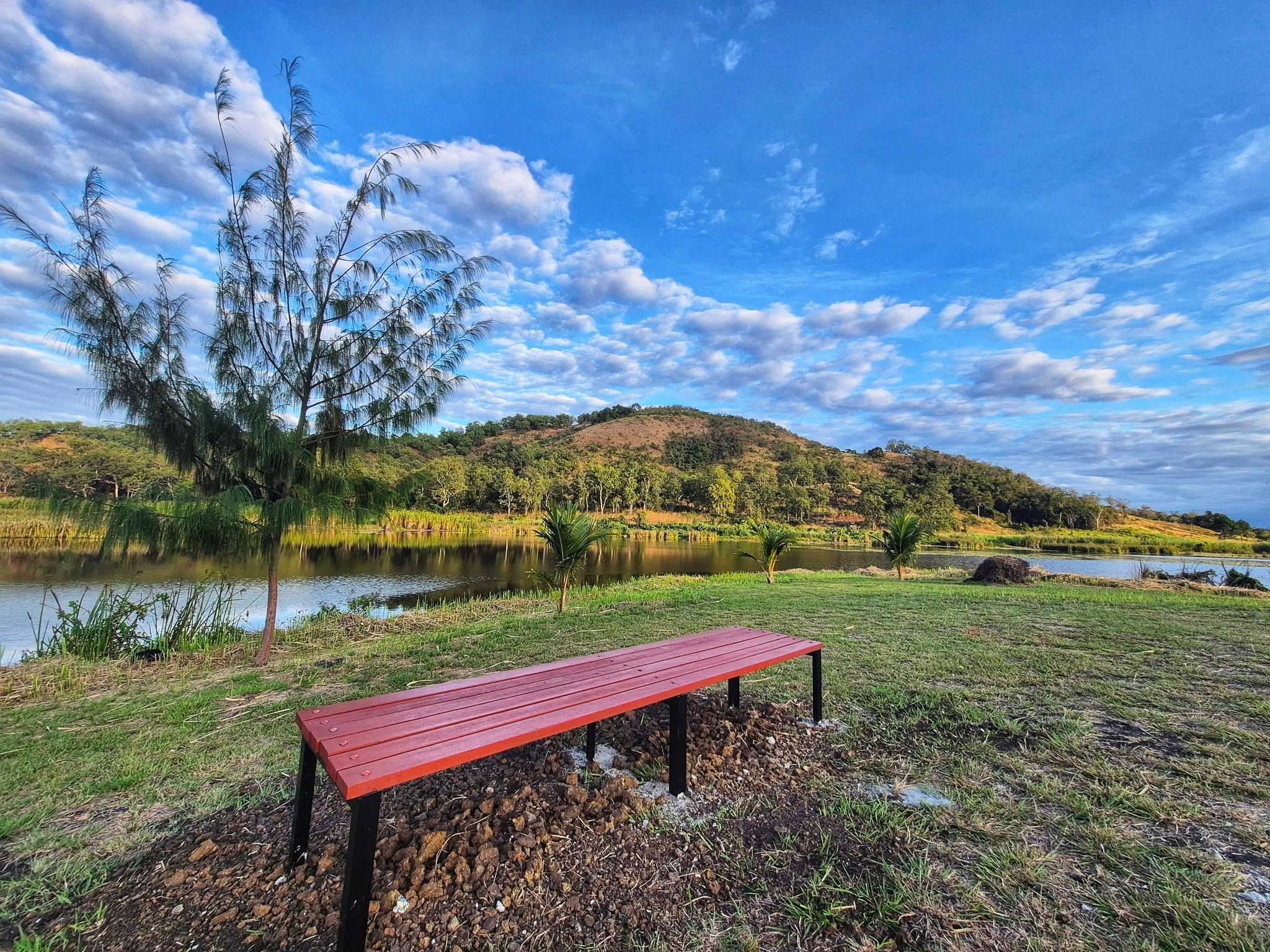 A simple red wooden bench on the grass facing a calm lake and rolling hills.